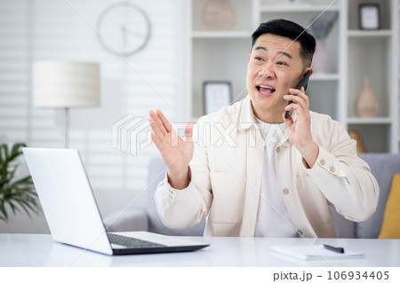 Mature asian man talking on the phone at home, man working in home office sitting at desk in home office, happy and smiling, consulting clients remotely. 106934405