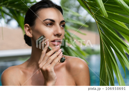 Beauty portrait, soft skin. Female model with natural make-up and healthy skin behind a green leafy plant. Portrait of a beautiful girl with nude nails, bare shoulders holding roller massager outdoor Beauty portrait, soft skin. Female model with natural make-up and healthy skin behind a green leafy plant. Portrait of a beautiful girl with nude nails, bare shoulders holding roller massager outdoor 106938691