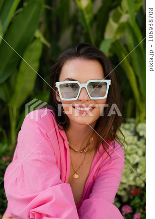 Young beautiful brunette in a pink shirt, neck jewelry, necklace, trendy sunglasses sits on a background of tropical leaves, laughs, smiles 106938708