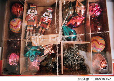 Cardboard gift box with colorful glittery Christmas tree glass toys. Vintage retro background. New Year's Eve 2024. Winter vacations ornament festive decor for home interior. Nutcracker, stars symbols Cardboard gift box with colorful glittery Christmas tree glass toys. Vintage retro background. New Year's Eve 2024. Winter vacations ornament festive decor for home interior. Nutcracker, stars symbols 106940452
