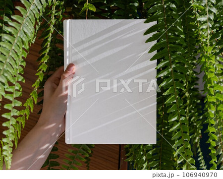 Hands holding book in hardcover mock up among green fern leaf plant Hands holding book in hardcover mock up among green fern leaf plant 106940970