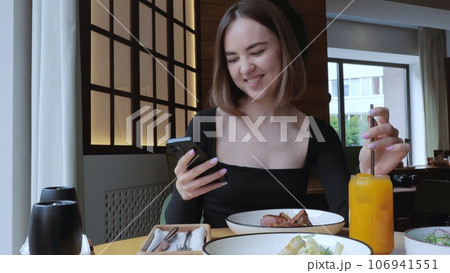The girl smiles while looking at the phone. A young woman is having dinner in a restaurant, drinking orange juice and eating a steak. Beautiful Woman has lunch in a cafe, lunch in a restaurant. 106941551