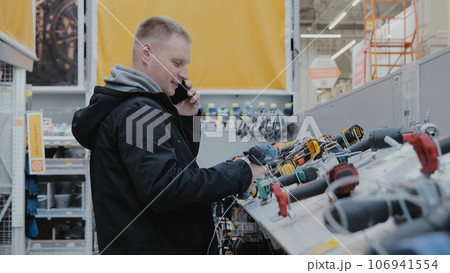 Buying a screwdriver at a hardware store. A man in a hardware store buys a power tool. Cordless power tools. Construction equipment supermarket Buying a screwdriver at a hardware store. A man in a hardware store buys a power tool. Cordless power tools. Construction equipment supermarket 106941554