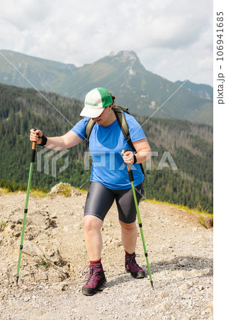 Happy woman hiker with backpack standing on the slope of mountain ridge against mountains. 30s Women holding hiking sticks. Travel and active lifestyle concept, outdoor activities Happy woman hiker with backpack standing on the slope of mountain ridge against mountains. 30s Women holding hiking sticks. Travel and active lifestyle concept, outdoor activities 106941685