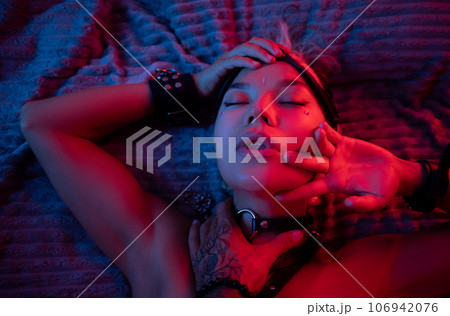 portrait of a sexy girl in a leather mask and bdsm accessories on a bed enjoying sex in orgasm with emotions on her face in neon light 106942076