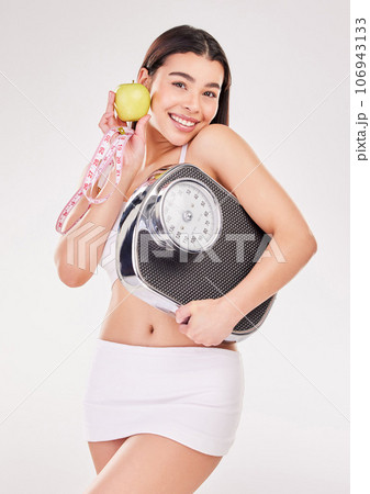 An apple a day.... Studio shot of an attractive young woman posing with an apple, measuring tape and weightscale against a grey background. An apple a day.... Studio shot of an attractive young woman posing with an apple, measuring tape and weightscale against a grey background. 106943133