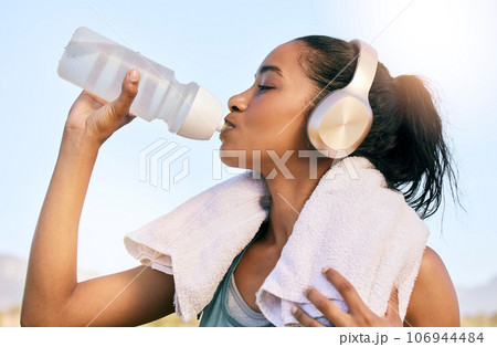 An active fit woman wearing wireless headphones drinking water from a bottle after exercising outdoors. Female athlete quenching thirst and cooling down with a towel after training workout outside An active fit woman wearing wireless headphones drinking water from a bottle after exercising outdoors. Female athlete quenching thirst and cooling down with a towel after training workout outside 106944484