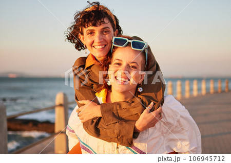 Lesbian, portrait and couple of friends at the beach in piggyback, support and happy for lgbtq love. Sunset, sea and smile of women or gay people on summer date with pride, freedom and gen z holiday 106945712