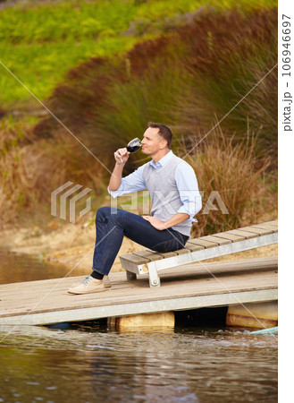 Thinking, river and a man drinking wine outdoor in nature on his farm for agriculture or sustainability. Water, glass and idea with a male person sitting on the pier to drink alcohol alone outside 106946697