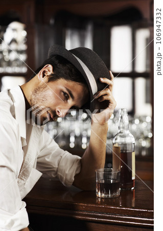 Portrait, whisky and a man sitting at the bar drinking alcohol in a glass while alone or feeling unhappy. Brandy, drink and beverage with a handsome young male pub customer at a wooden counter Portrait, whisky and a man sitting at the bar drinking alcohol in a glass while alone or feeling unhappy. Brandy, drink and beverage with a handsome young male pub customer at a wooden counter 106947332