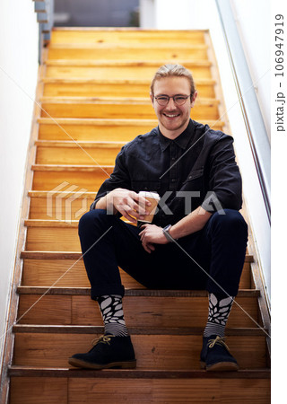 I need coffee to recharge. Full length portrait of a handsome young businessman sitting on the office staircase and holding a cup of coffee. 106947919
