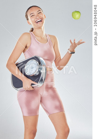 Happy woman, diet and scale to lose weight with apple, fitness goals or progress in studio. Portrait, laughing or female sports model throw fruit for healthy nutrition, weightloss results or wellness 106948048