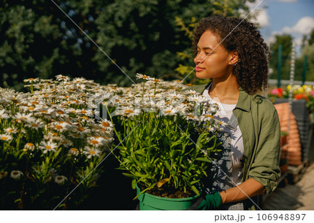 Pretty woman gardener holding flower pot while standing on greenhouse yard background 106948897