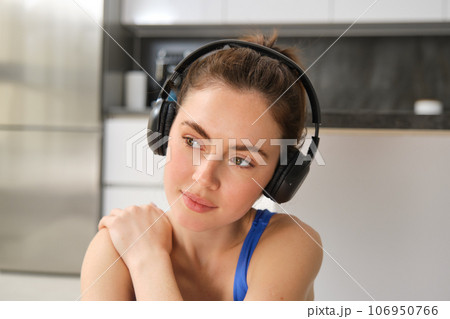 Close up portrait of brunette fitness girl, listens music in wireless headphones, smiles at camera, does workout alone at home, sits on floor and exercises 106950766