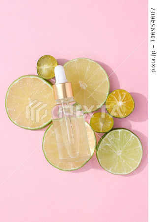 A transparent dropper bottle placed on slices of kumquat and lime on a pink background. Minimal advertising scene for cosmetic and product with ingredient from natural essential 106954607