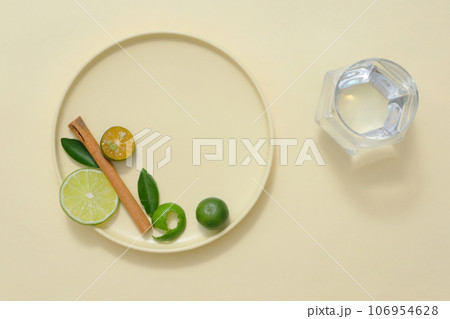 A round ceramic dishes containing slices of lime and kumquat, green leaves and dried cinnamon stick decorated with glass cup of water. Space for design 106954628