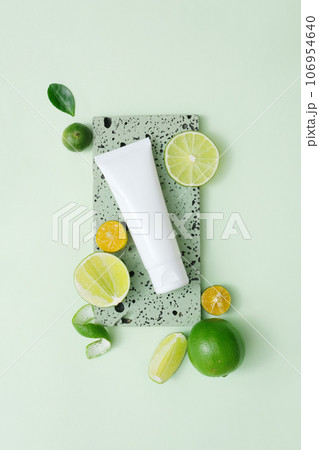 A white plastic tube with facial moisturizer cream or facial cleanser decorated with slices of lime and kumquat on gray brick podium on pastel background 106954640
