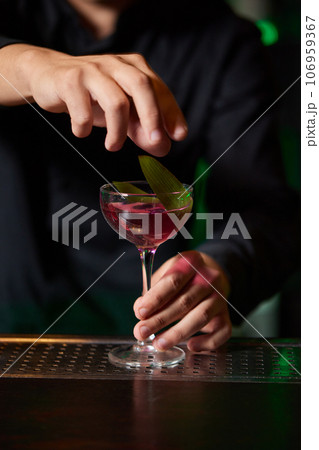 Barman in shirt and apron making an alcoholic drink with ice in a cocktail glass. 106959367