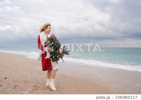 Blond woman holding Christmas tree by the sea. Christmas portrait of a happy woman walking along the beach and holding a Christmas tree in her hands. Dressed in a red coat, white dress. 106960327