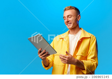Portrait of young handsome smiling guy, student dressed in bright fashion outfit holding tablet isolated on blue background. 106965145