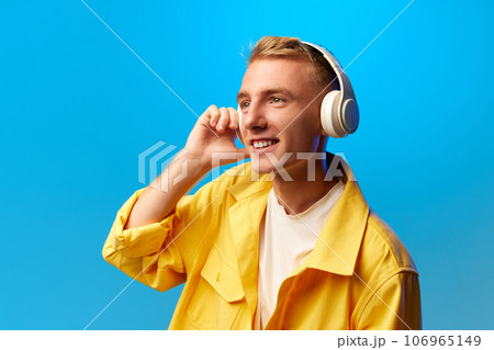Portrait of young handsome smiling guy, student in bright fashion outfit and listening misic in headphones isolated on blue background. 106965149