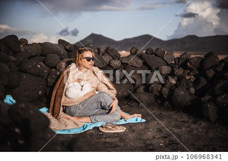 Mother breast feeding his infant baby boy son on black sandy volcanic beach of Janubio on Lanzarote island, Spain, enjoing dramatic volcanic landscape on windy overcast day. Travel with kids concept. 106968341
