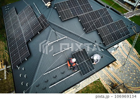 Engineers building photovoltaic solar module station on roof of house. Men electricians in helmets installing solar panel system outdoors. Concept of alternative and renewable energy. Aerial view. Engineers building photovoltaic solar module station on roof of house. Men electricians in helmets installing solar panel system outdoors. Concept of alternative and renewable energy. Aerial view. 106969134