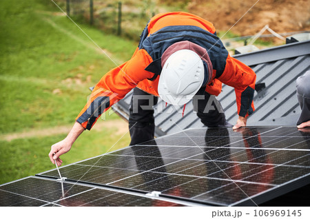 Builders installing photovoltaic solar panels on roof of house. Men engineers in helmets building solar module system with help of hex key. Concept of alternative, renewable energy. 106969145