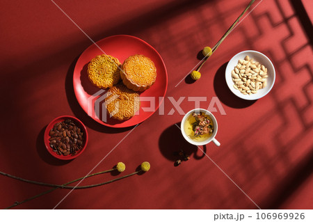 Layout of a traditional tea table party on Mid-autumn festival occasion. Delicious mooncakes on ceramic dish, tea cup and dry types of seed decorated on red background with window shadow 106969926