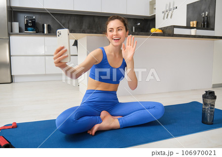Vlogger does sports, records her workout training from home on smartphone camera, posing for selfie inside her house, sits on rubber yoga mat in blue leggings and sportsbra 106970021