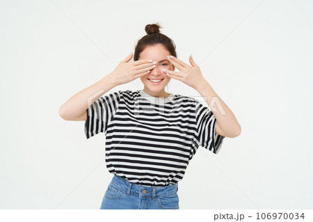 Portrait of girl waiting for surprise, covers eyes with hands, peeking through fingers, stands over white background 106970034