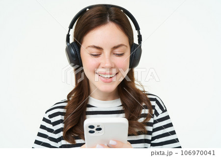 Image of young woman in headphones, using smartphone and laughing, watching video on mobile phone, listens to music on streaming service app, white background Image of young woman in headphones, using smartphone and laughing, watching video on mobile phone, listens to music on streaming service app, white background 106970041