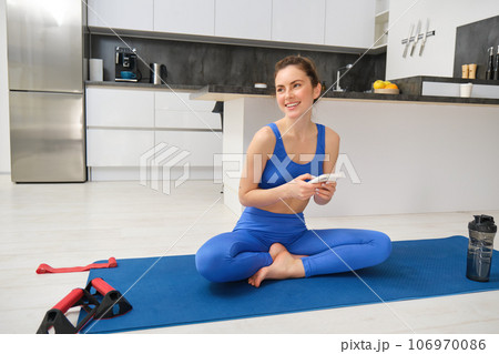 Sport and wellbeing. Young sportswoman does exercises at home, sits in yoga pose, holds smartphone, follows online training videos during her workout indoors Sport and wellbeing. Young sportswoman does exercises at home, sits in yoga pose, holds smartphone, follows online training videos during her workout indoors 106970086