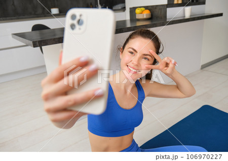 Portrait of girl takes selfie on smartphone while does her workout training. Young woman does her exercises at home, takes selfie for sports blog, sits in living room 106970227