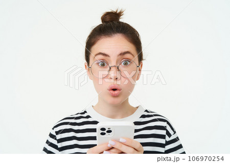 Image of girl in glasses, holding smartphone and looking surprised, amazed by smth on mobile phone, stands over white background 106970254