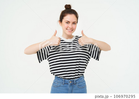 Portrait of cute girl, student shows thumbs up, recommends product, approves choice, likes item, stands over white background Portrait of cute girl, student shows thumbs up, recommends product, approves choice, likes item, stands over white background 106970295