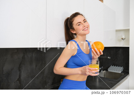Indoor shot of woman after workout, standing in kitchen with fresh juice and an orange, drinking it 106970307