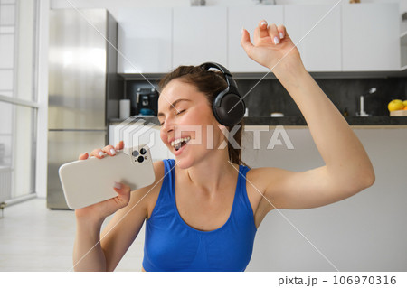 Happy young girl doing fitness, singing and having fun, listening music in headphones, holding smartphone, workout in living room Happy young girl doing fitness, singing and having fun, listening music in headphones, holding smartphone, workout in living room 106970316