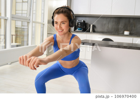 Portrait of beautiful young woman in blue sportswear, doing squats and stretching arms, workout at home, aerobics exercises, listening to music in wireless headphones 106970400