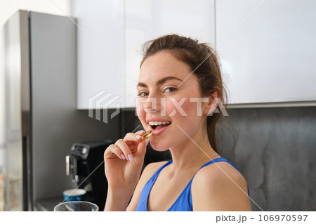 Close up portrait of smiling sportswoman, fitness girl taking vitamins, fish oil pill, using dietary supplements for strong health and fit body, standing in kitchen Close up portrait of smiling sportswoman, fitness girl taking vitamins, fish oil pill, using dietary supplements for strong health and fit body, standing in kitchen 106970597
