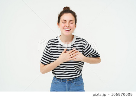 Portrait of happy, smiling young woman stands with eyes closed, holds hands on heart, laughs, remember something funny, isolated over white background 106970609