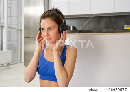 Image of fitness girl doing sports at home, puts on wireless headphones for music during workout training session, wearing blue sportsbra 106970677