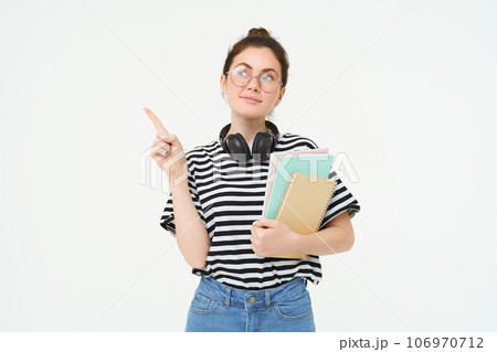 Image of smart young woman in glasses, student carries her notebooks and planners, points left with thinking face, making choice, deciding on smth, white background 106970712