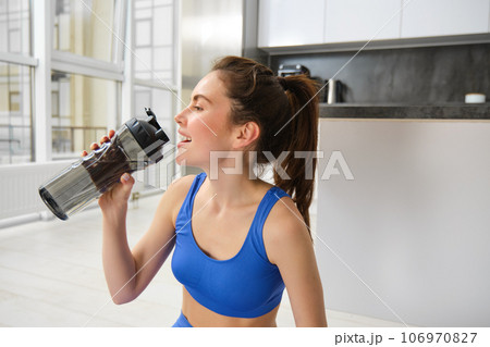 Portrait of sportswoman drinking water during workout session in living room, does her fitness training exercises at home Portrait of sportswoman drinking water during workout session in living room, does her fitness training exercises at home 106970827