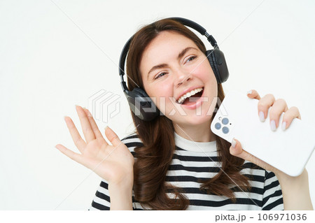 Technology and social media. Portrait of happy young woman singing karaoke, playing music app on smartphone, wearing wireless headphones, standing over white background 106971036
