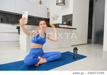 Portrait of active girl, vlogger does sports, records her workout training from home on smartphone camera, posing for selfie inside her house, sits on rubber yoga mat in blue leggings and sportsbra Portrait of active girl, vlogger does sports, records her workout training from home on smartphone camera, posing for selfie inside her house, sits on rubber yoga mat in blue leggings and sportsbra 106971099