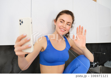 Portrait of young fitness girl, social media blogger, live streaming, saying hello at smartphone, waving hand and smiling, wearing activewear 106971121