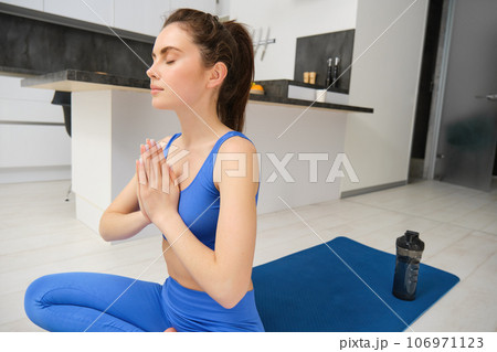 Sport and recreation. Close up portrait of beautiful girl does yoga at home, clasps her hands and meditates, sits on rubber mat in kitchen, makes asana, relaxing Sport and recreation. Close up portrait of beautiful girl does yoga at home, clasps her hands and meditates, sits on rubber mat in kitchen, makes asana, relaxing 106971123