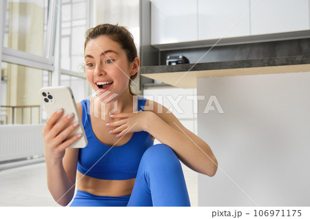 Indoor shot of surprised, happy young sportswoman looking at smartphone with smile and excitement, reading great news on phone message, sitting in blue sportsbra and leggings 106971175