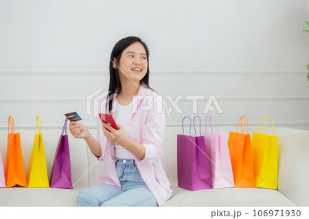 Young asian woman sitting on sofa using smart phone shopping online with credit card while paper bag 106971930